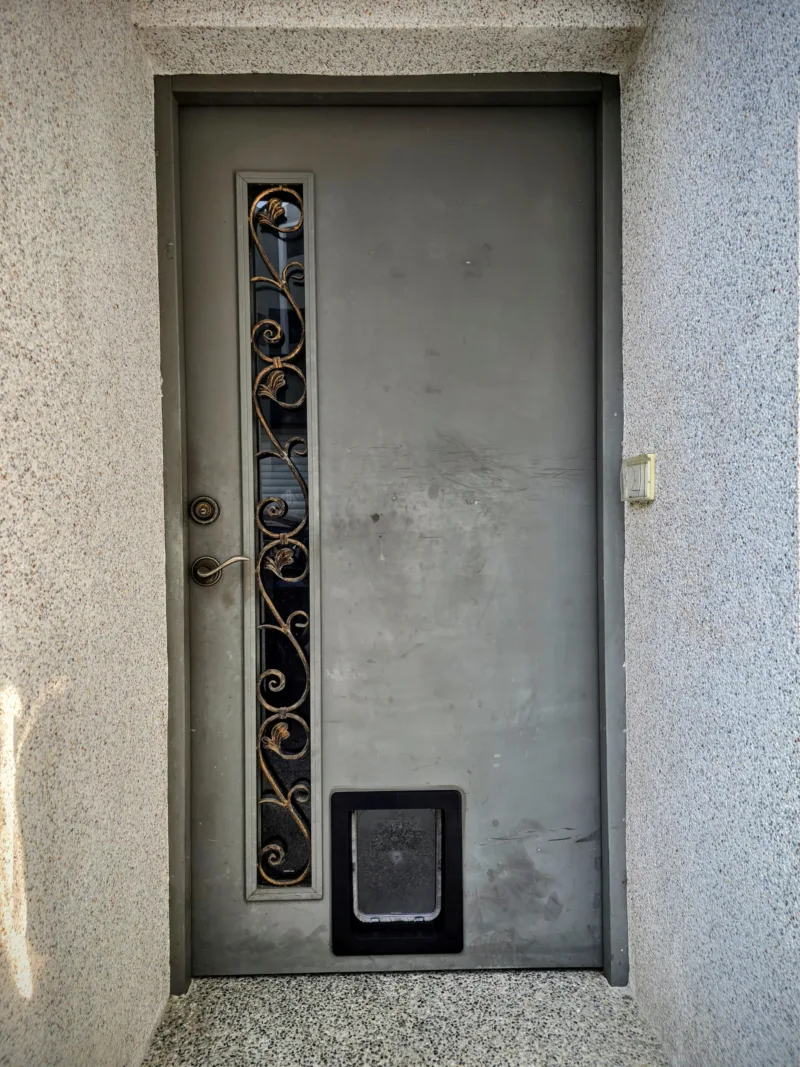 從室外拍攝的灰色車庫小門完工照。門板下方正中間偏左處(避開左側鍛鐵裝飾玻璃)已精準切割並安裝好黑色的寵物貓門。安裝邊緣緊密、毫無破綻,維持了原本大門的完整性與美觀,右側牆面為洗石子材質,呈現出與原有建築融為一體的實用設計。